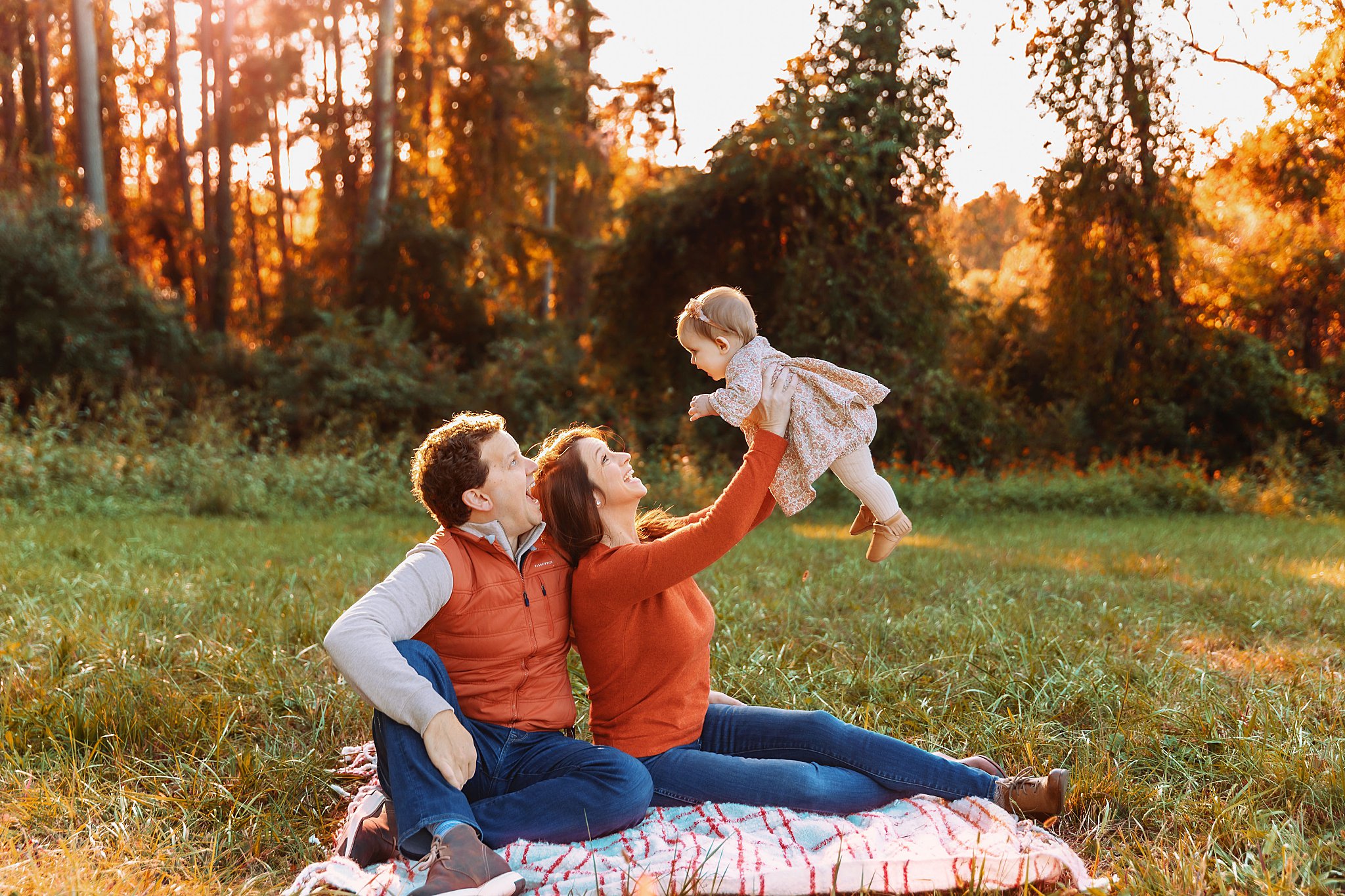 parents sit on blanket in woods as they hold baby in the air by Charlottesville photographer
