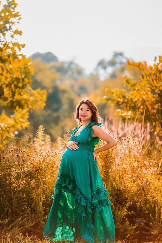 warm sun shines on woman and flowers as she holds her pregnant stomach by Amy Yang Photography