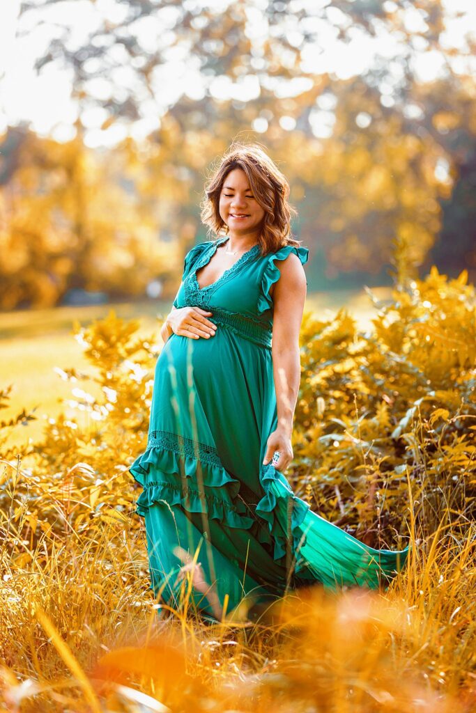 woman in green dress smiles as she holds her pregnant belly during outdoor maternity session