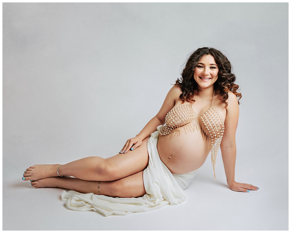 mom-to-be sits on floor leaning on one arm in beaded top and flowing skirt for luxe studio maternity session