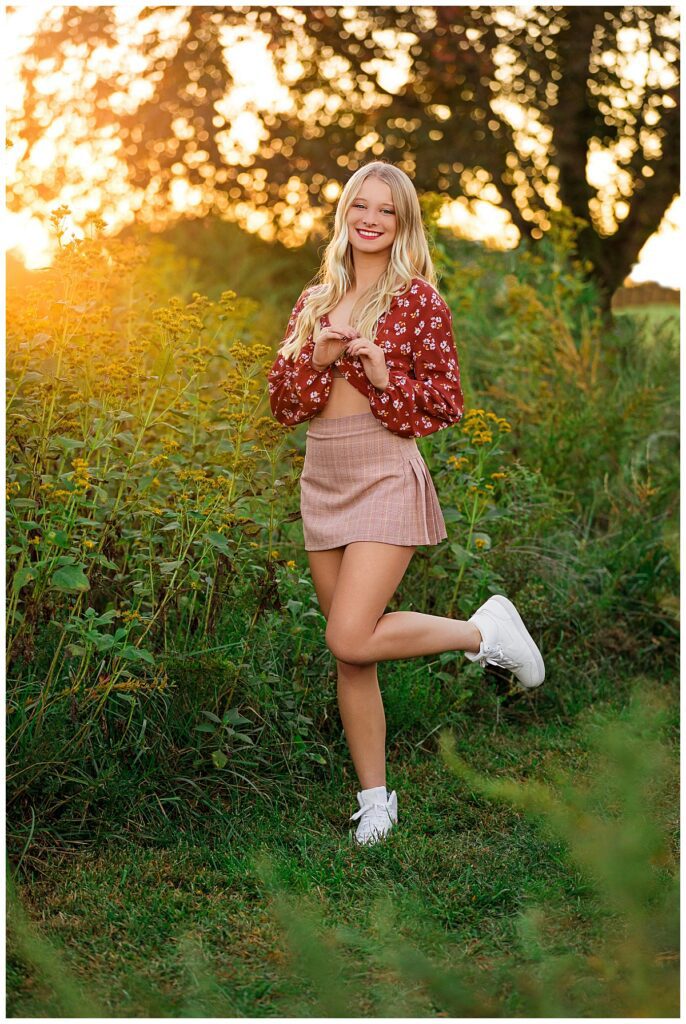 High school senior smiling in a wildflower field during golden hour in a floral top and skirt, showing what to wear for senior pictures with a Senior Photographer in Charlottesville, VA.
