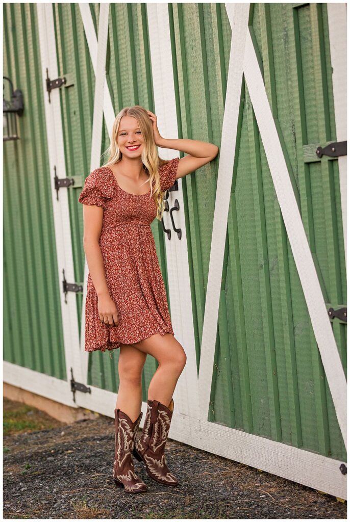 High school senior in a red floral dress and cowboy boots posing in front of a green barn.