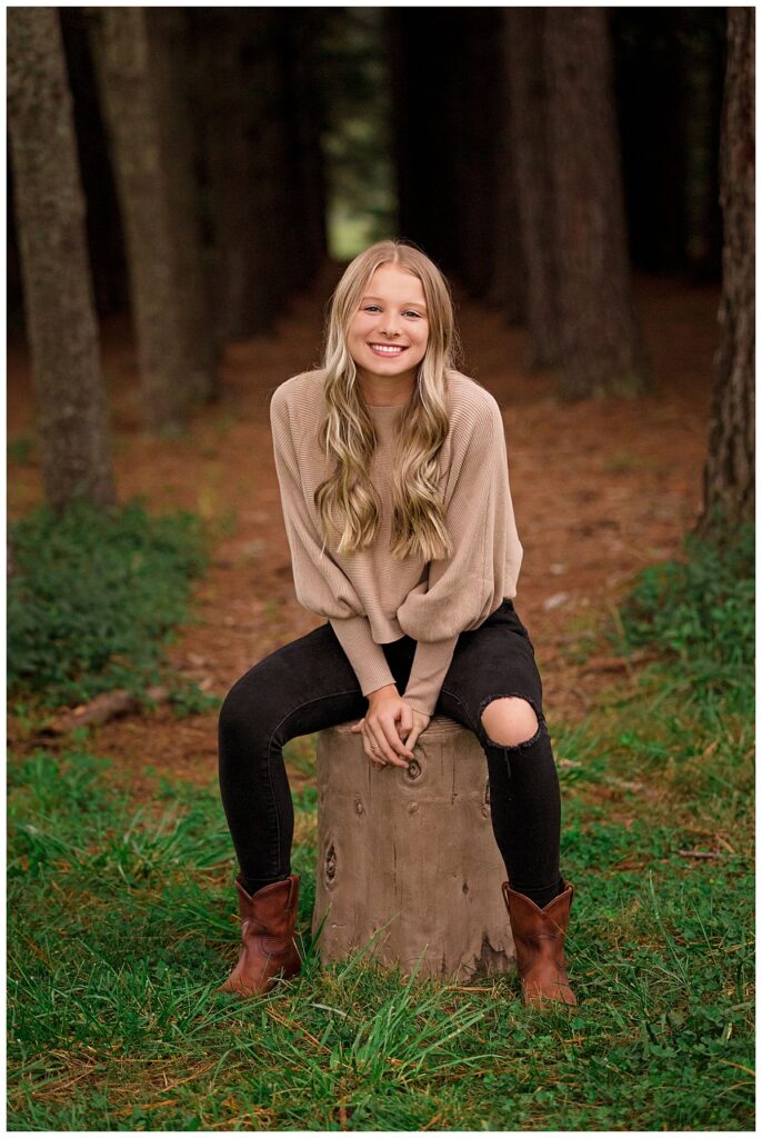 High school senior sitting on a tree stump in a wooded area, smiling during an outdoor senior portrait session.