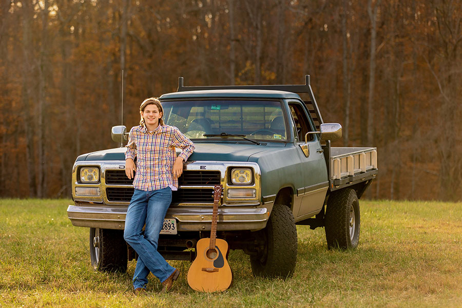 High school senior leaning against a vintage pickup truck with an acoustic guitar in an open field for a personalized outdoor senior portrait session.