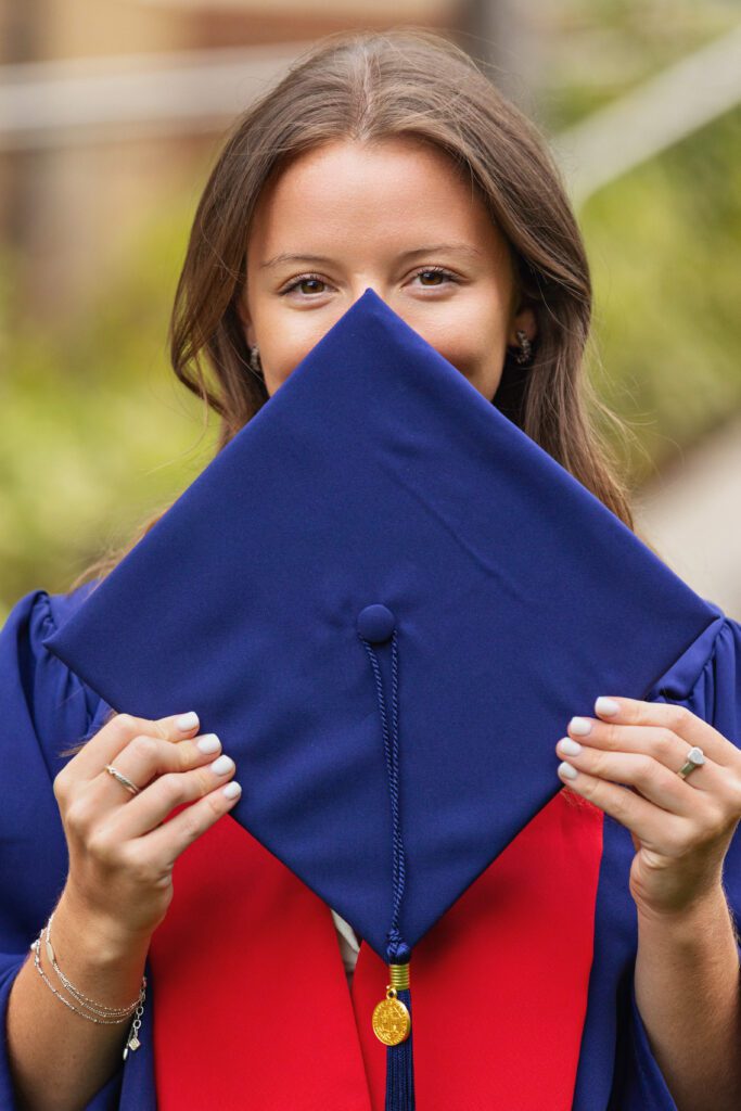Graduate holding graduation cap over her face while smiling during a college graduation session by a Charlottesville Senior Photographer. Learn when to book senior pictures for the best portraits.