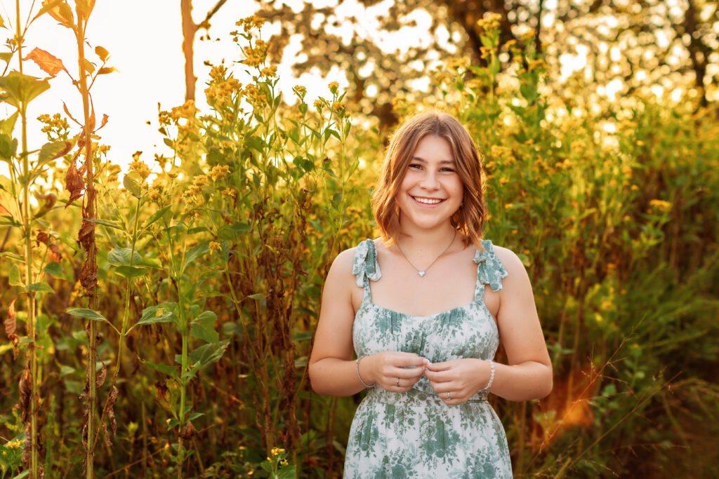 Smiling high school senior in a floral dress standing among wildflowers during golden hour, showcasing the best time of day for senior pictures with a Charlottesville Senior Photographer.