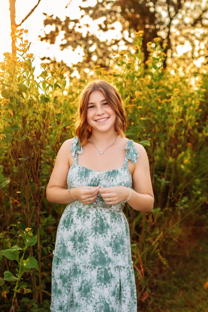 Smiling high school senior in a floral dress standing in a field surrounded by warm evening light and wildflowers during an outdoor senior portrait session.
