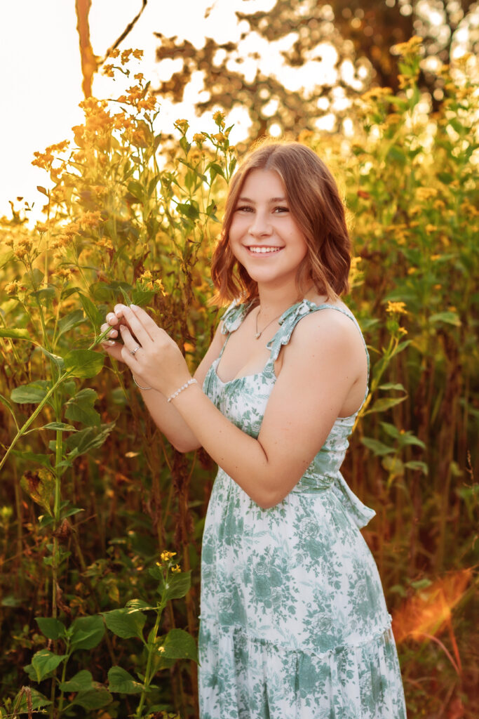 High school senior gently holding wildflowers while smiling during a golden hour senior portrait session in a blooming field near Charlottesville, Virginia.