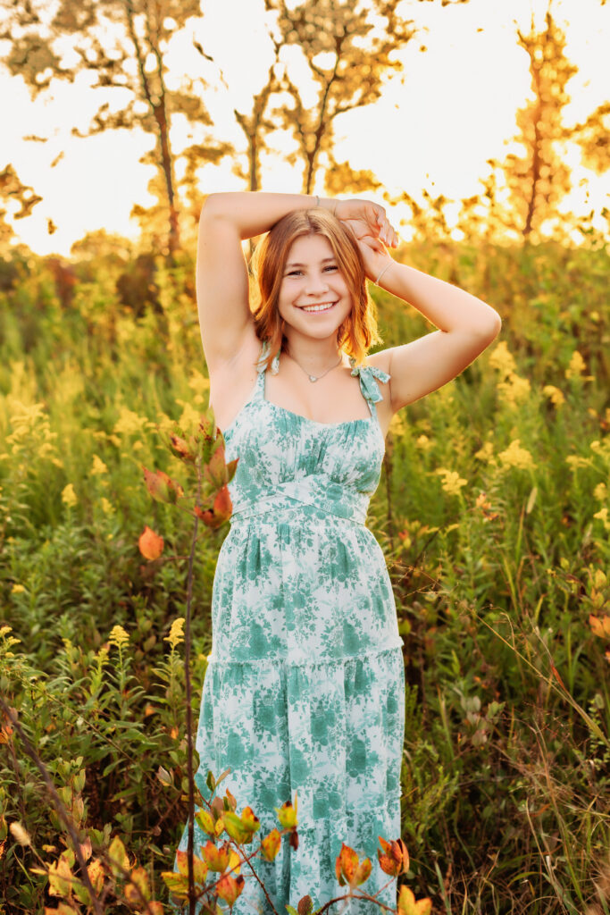 Senior portrait of a smiling high school senior in a light blue floral dress posing in a sunlit field, highlighting the best time of day for senior pictures by a Charlottesville Senior Photographer.