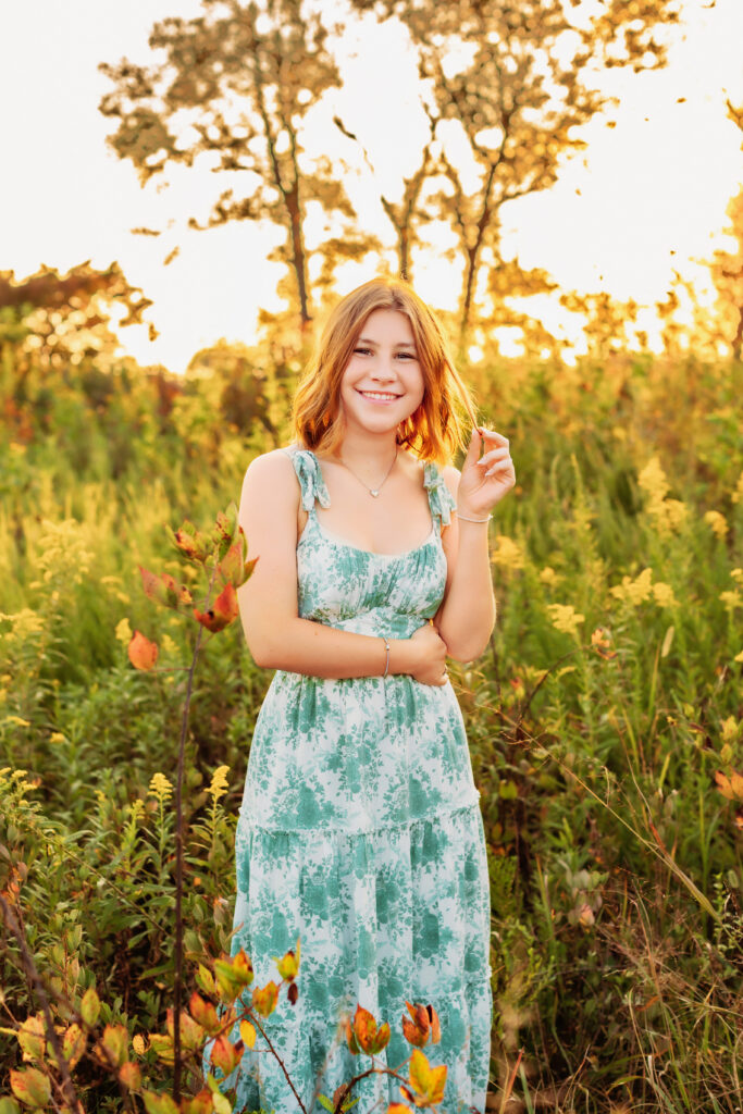 High school senior smiling in a golden hour meadow wearing a floral dress, with a Charlottesville Senior Photographer.