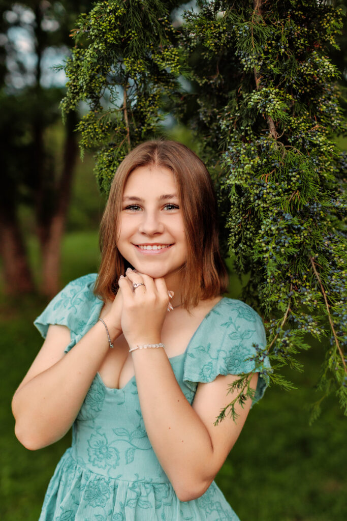 Close-up portrait of a smiling high school senior in a light blue dress beneath evergreen branches during an outdoor senior photo session in Richmond, VA.