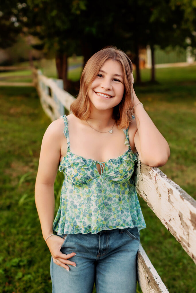 Smiling high school senior leaning against a white fence during an outdoor session.