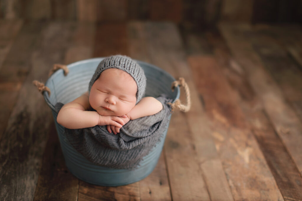 Luxury newborn photography portrait of a sleeping baby in a blue bucket with a gray knit bonnet and wrap in Charlottesville, VA.