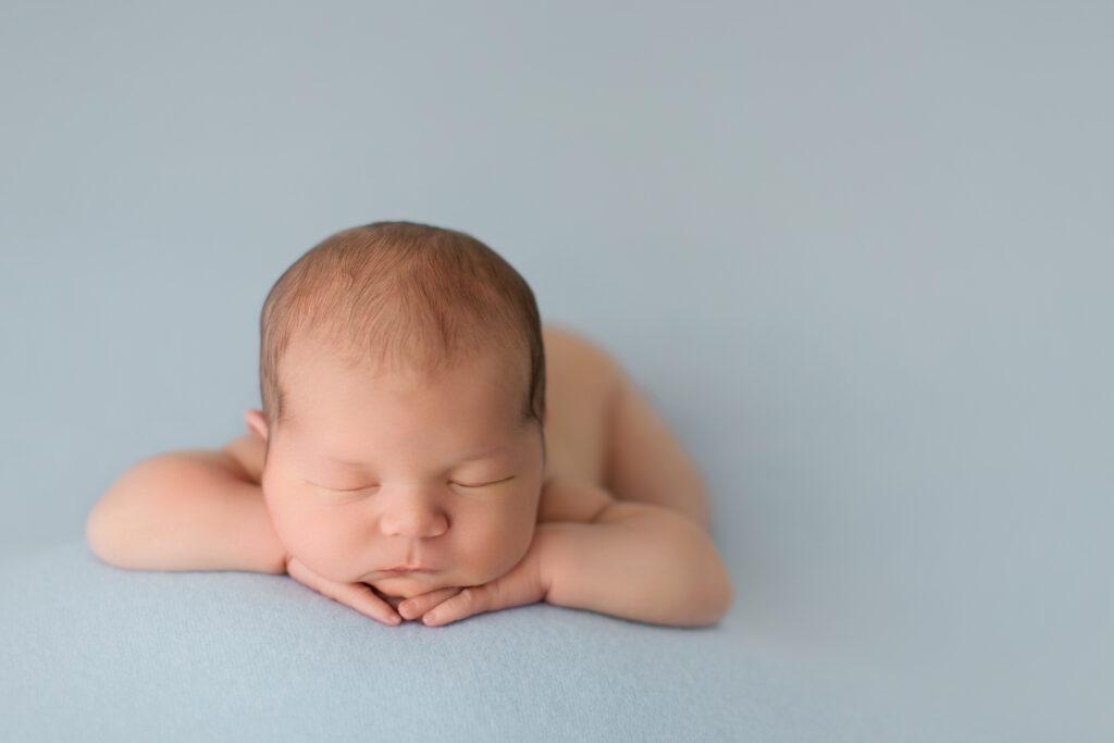 Peaceful newborn resting on folded hands against a soft blue backdrop during a luxury newborn photography session with Amy Yang Photography.