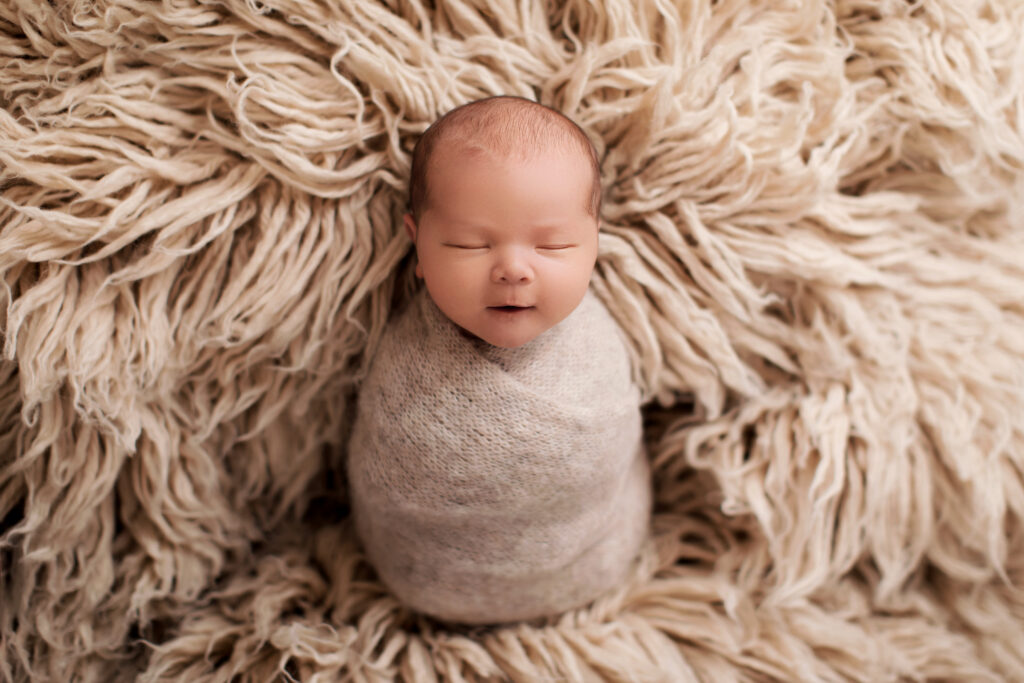 Wrapped sleeping newborn surrounded by a textured cream blanket in a timeless luxury newborn photography portrait.