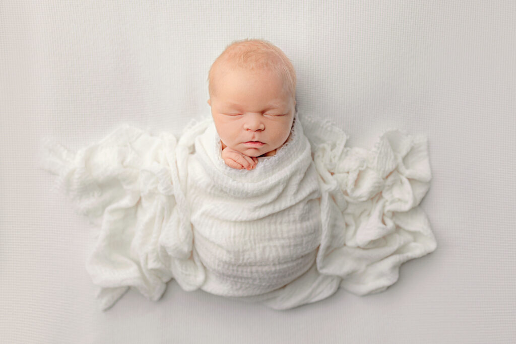 Peacefully sleeping newborn wrapped in white fabric photographed by a newborn photographer Crozet VA in a bright studio setting.