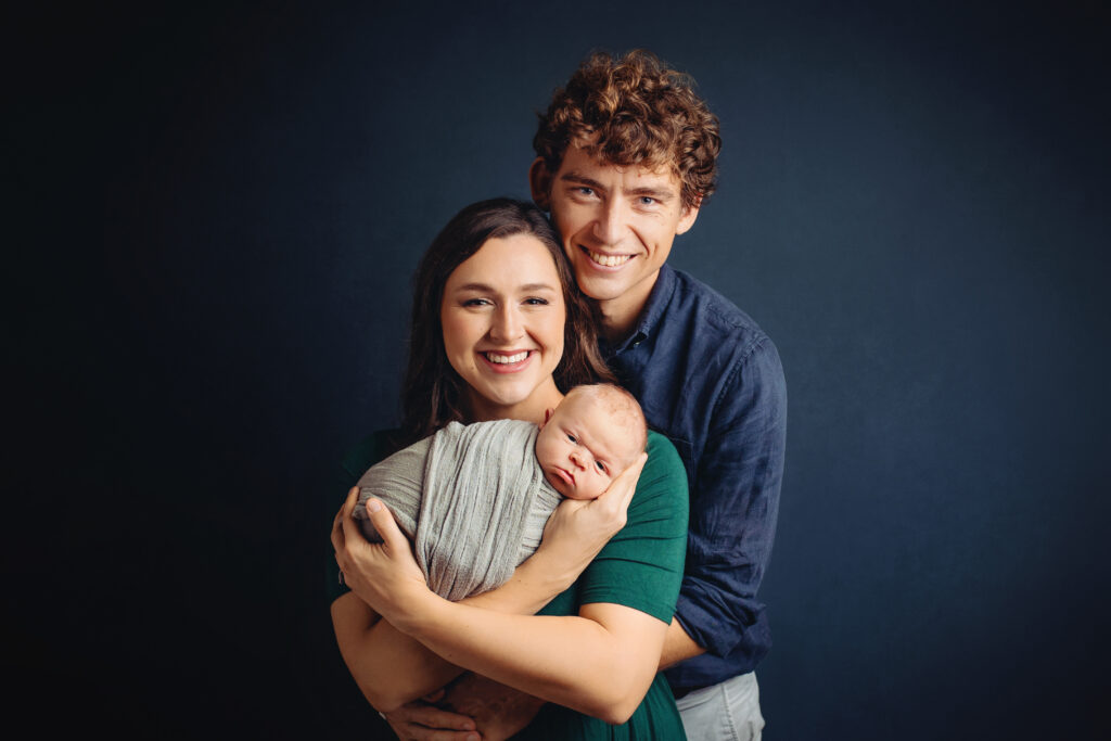 Newborn photographer Ivy VA capturing a smiling family holding their wrapped newborn during a studio portrait session.
