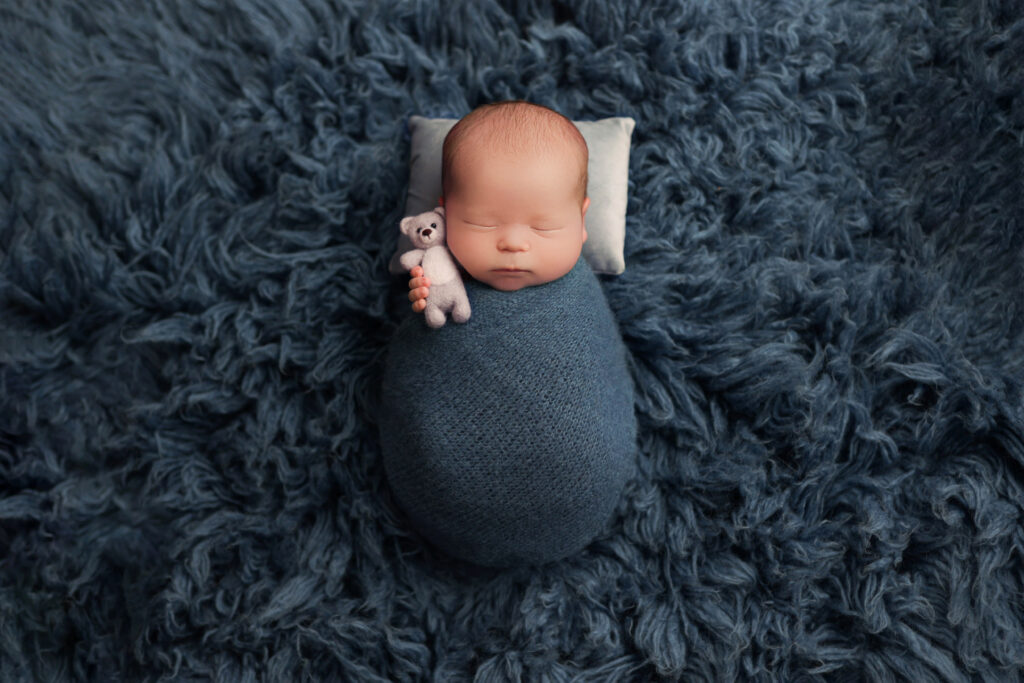 Newborn photos as home decor of a wrapped baby holding a small teddy bear on a blue blanket captured by Amy Yang Photography.