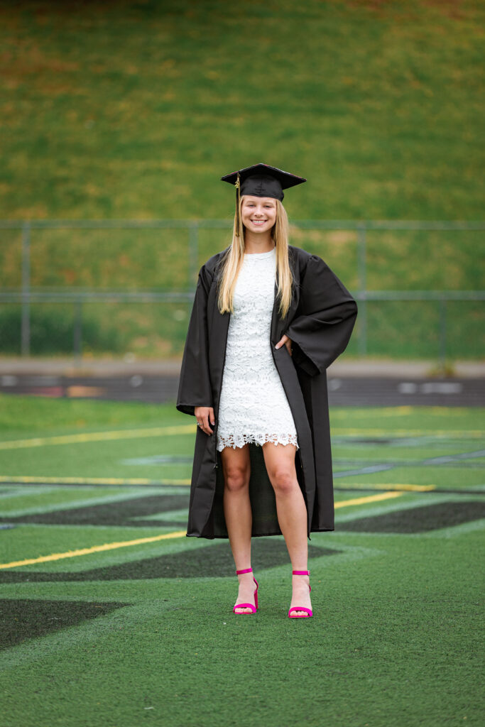 High school senior in a graduation cap and gown posing on a football field to celebrate graduation milestones.