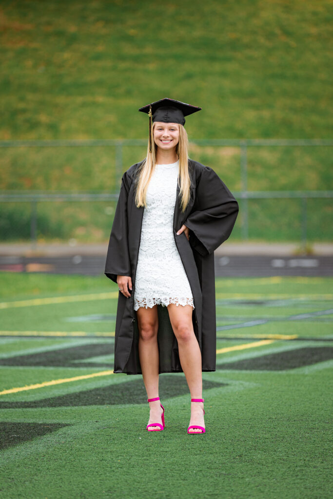 Smiling graduate wearing a black cap and gown over a white lace dress on a football field during senior graduation portraits.