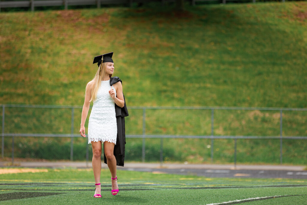 High school senior in a white dress and graduation cap walking across a football field, captured by a senior photographer in Richmond, VA.
