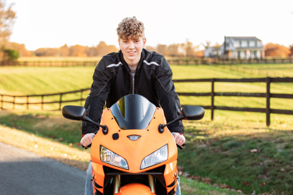 High school senior leaning over an orange sport motorcycle during an outdoor senior portrait session with a scenic countryside backdrop.