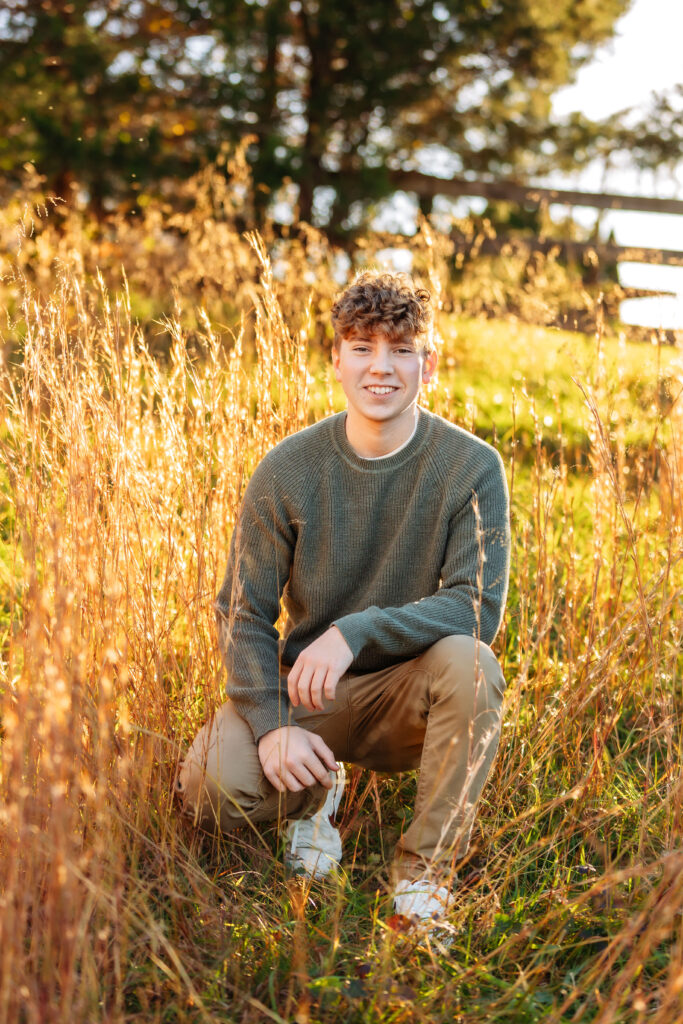 High school senior kneeling in tall golden grass during a relaxed outdoor senior portrait session in Charlottesville, Virginia.