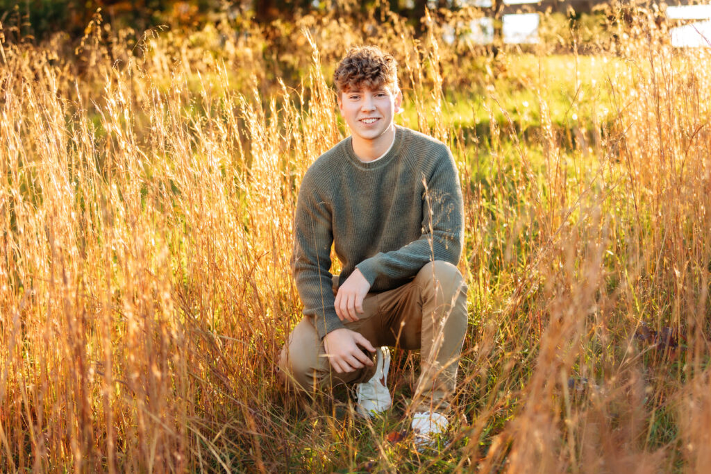 Smiling high school senior kneeling in tall golden grass during golden hour, capturing natural senior pictures for guys with a Charlottesville Senior Photographer.