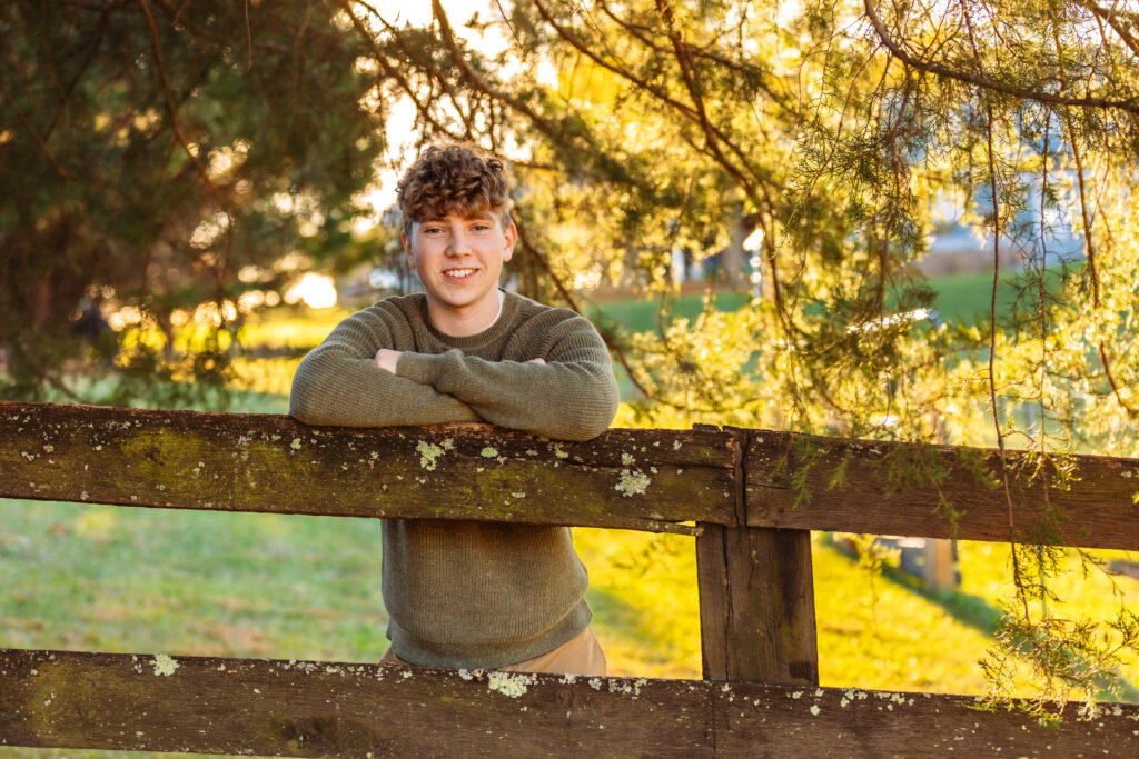 High school senior smiling while leaning on a rustic wooden fence during a golden hour outdoor portrait session.