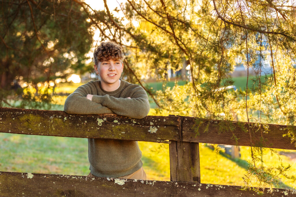 Relaxed high school senior leaning on a wooden fence beneath evergreen branches during a fall senior photo session in Charlottesville, VA.