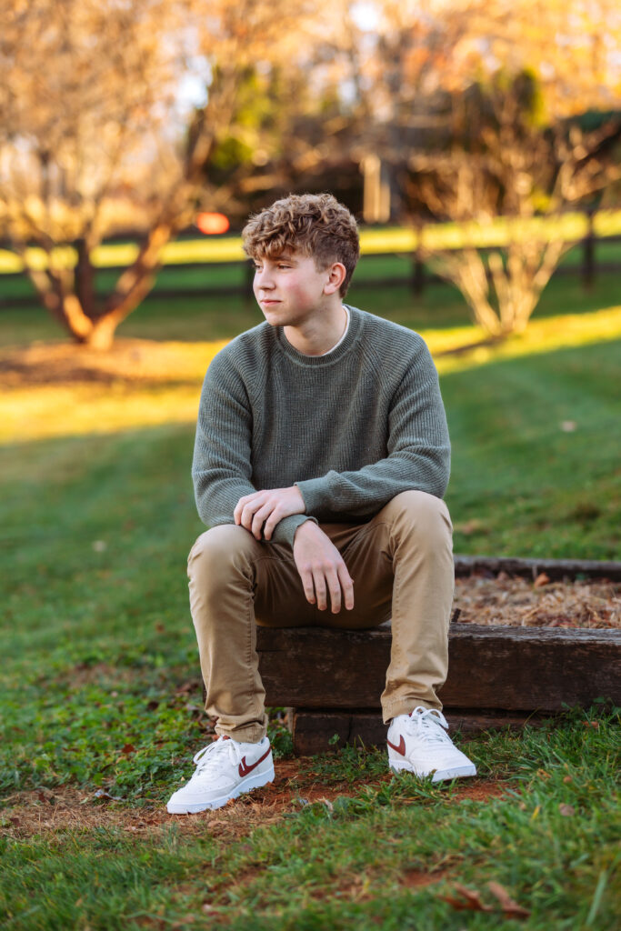 High school senior sitting outdoors in a green sweater and khaki pants surrounded by warm autumn colors during a natural light portrait session.