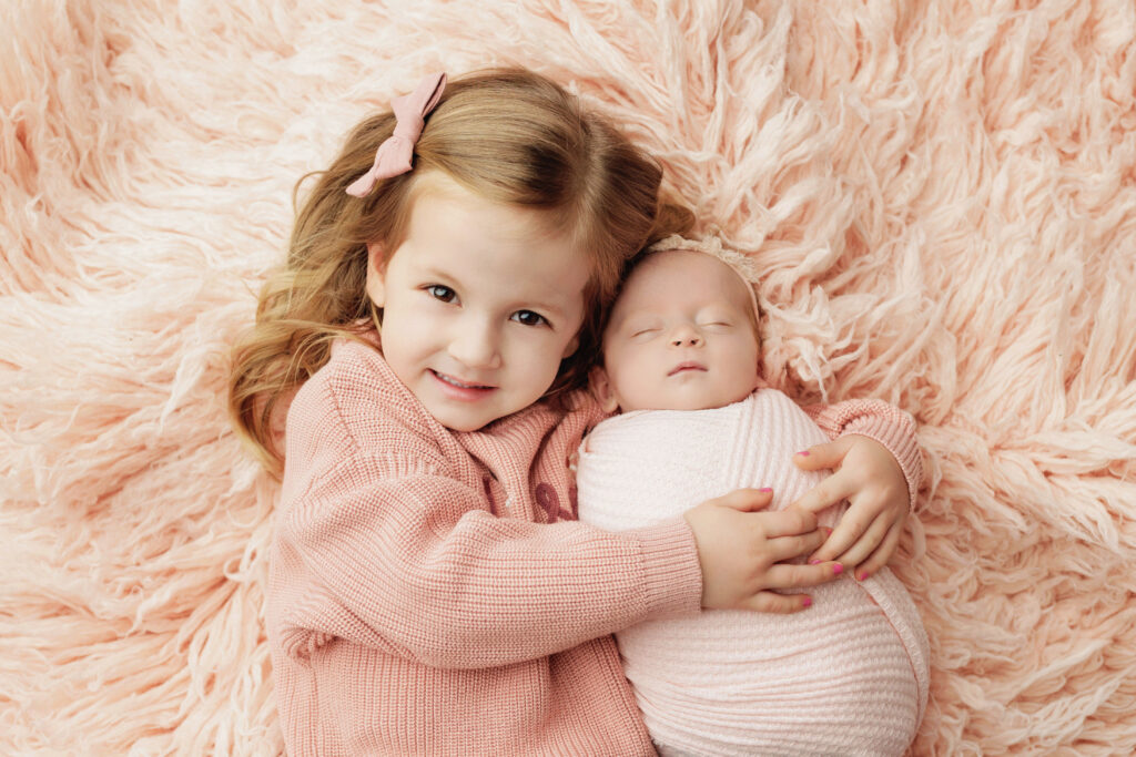 Big sister cuddling her newborn sibling on a soft pink blanket illustrating why print newborn photos.