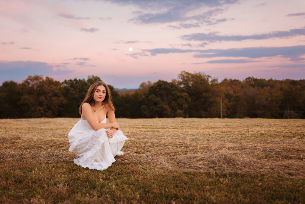 High school senior in a white dress crouching in a harvested field beneath a pastel sunset sky during an outdoor senior photography session.
