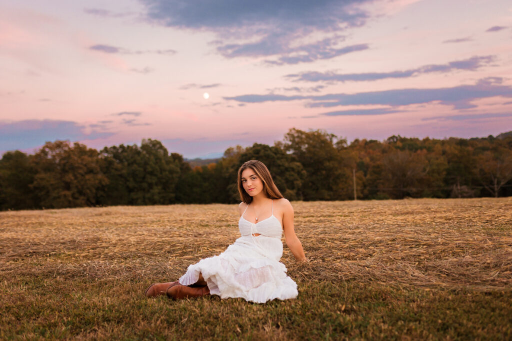 High school senior in a flowing white dress sitting in an open field at sunset with a colorful sky during an outdoor senior portrait session by Amy Yang Photography.