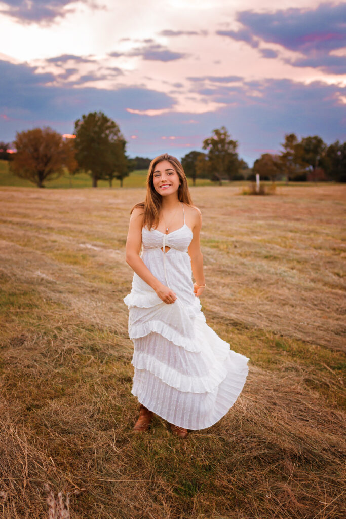 Smiling high school senior in a white dress standing in an open field beneath a colorful evening sky during a golden hour senior portrait session by Amy Yang Photography.