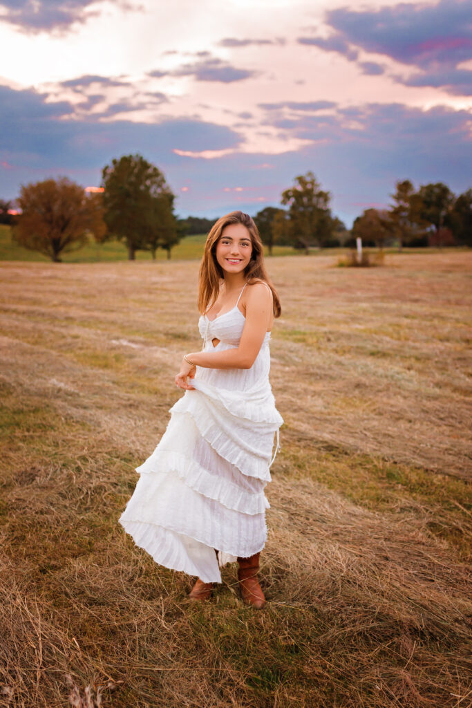 High school senior in a white dress walking through a scenic field at sunset, photographed by a professional senior photographer.