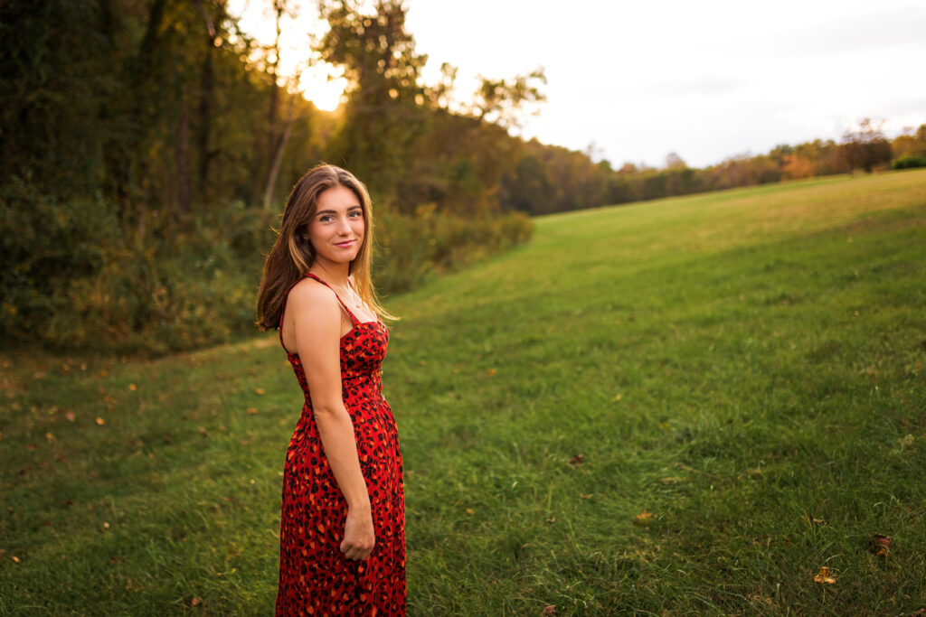 Smiling high school senior wearing a red patterned dress walking through a grassy field during a session with a professional senior photographer.