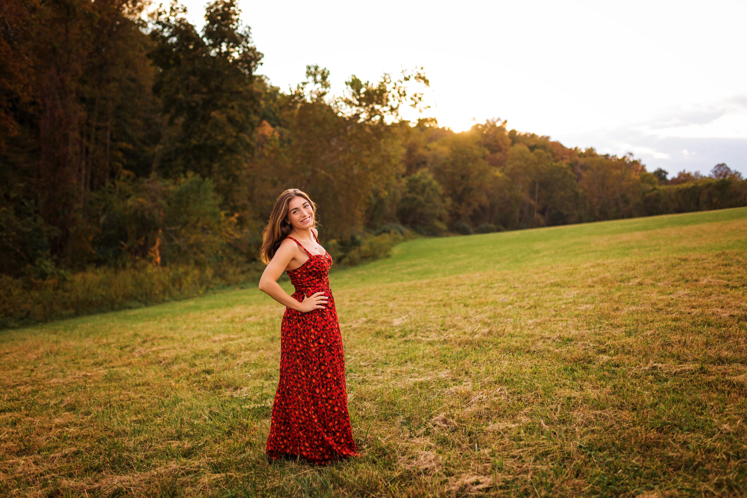 High school senior in a red maxi dress posing in an open field at golden hour with trees and rolling hills in the background, photographed by Amy Yang Photography.