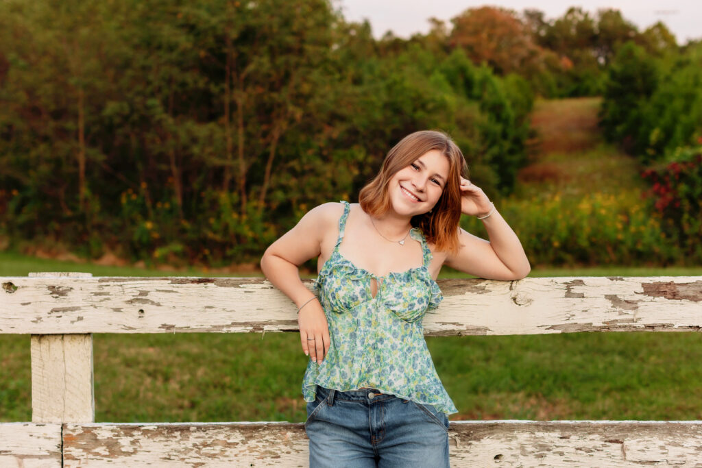 Smiling high school senior leaning against a rustic white fence during a senior photos session at sunset in Charlottesville, VA.
