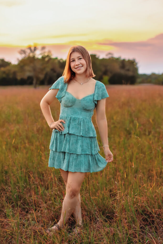 Smiling high school senior in a teal dress posing in a meadow at golden hour, showcasing the variety of a multiple locations senior photos session.