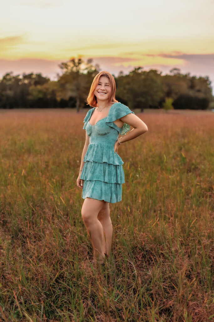 High school senior wearing a teal dress standing in a golden field at sunset during a multiple locations senior photos session.