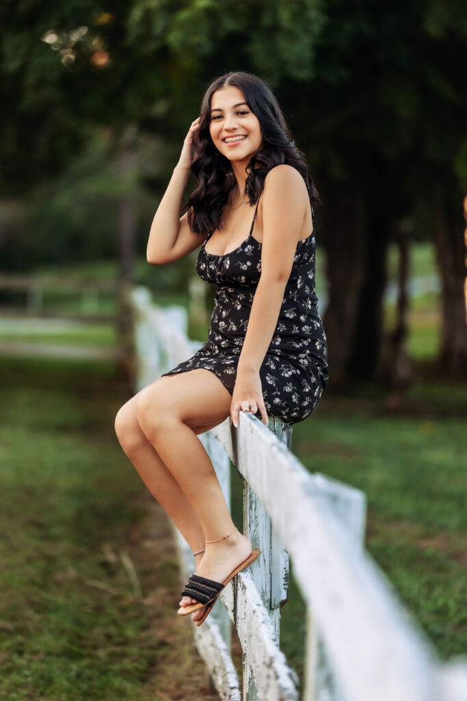 High school senior in a black floral dress leaning against a white fence on a scenic farm property during a Charlottesville, VA senior photo session.