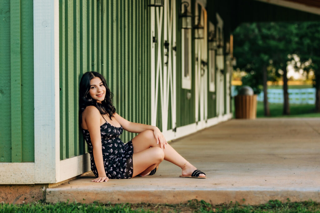 High school senior in a black floral dress sitting along a green barn during an outdoor senior portrait session in Charlottesville, VA.