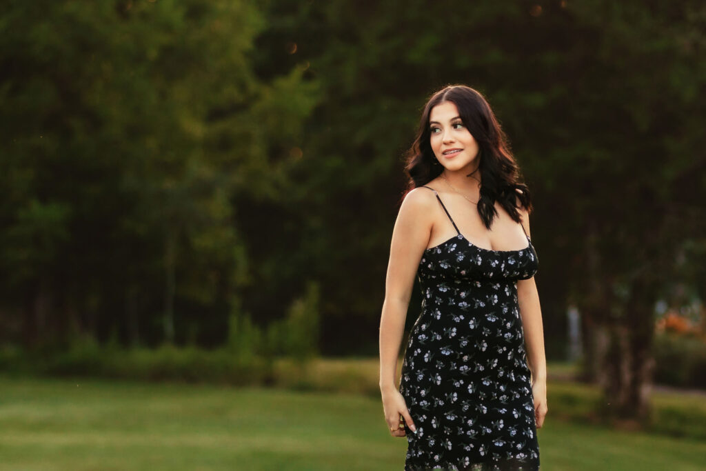 High school senior wearing a black floral dress standing in a grassy field at golden hour during an outdoor senior portrait session.